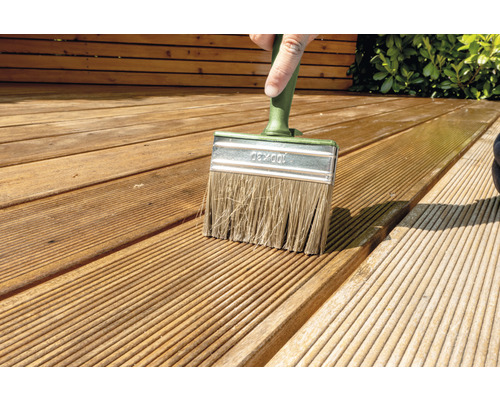 Een persoon schildert een houten terras met een platte kwast.