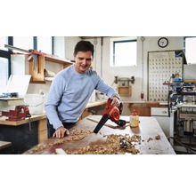 Een man gebruikt een accu bladblazer om een werkbank schoon te maken van houtsnippers in een werkplaats.