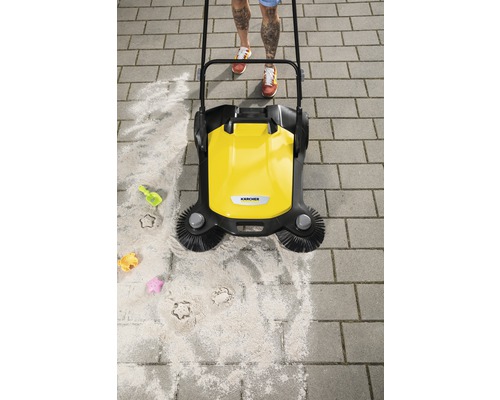 Gele veegmachine in gebruik op bestrating met zand en kinderspeelgoed.