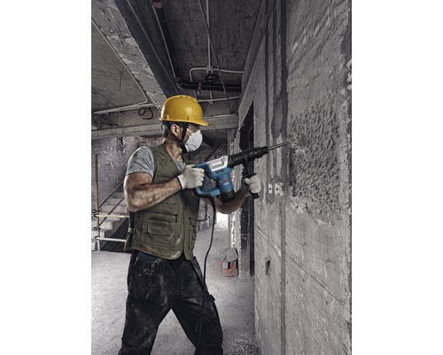Een bouwvakker met veiligheidshelm en stofmasker gebruikt een boorhamer op een betonnen muur.