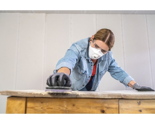 Vrouw schuurt houten plank met schuurblok, beschermd met handschoenen, veiligheidsbril en ademhalingsmasker.