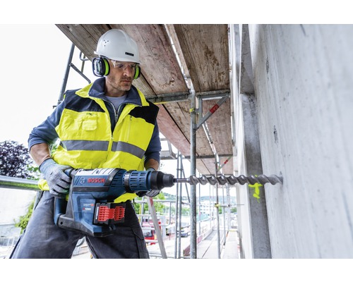Bouwvakker boort met een boorhamer een gat in een betonnen muur op een steiger. Hij draagt beschermende kleding en een helm.