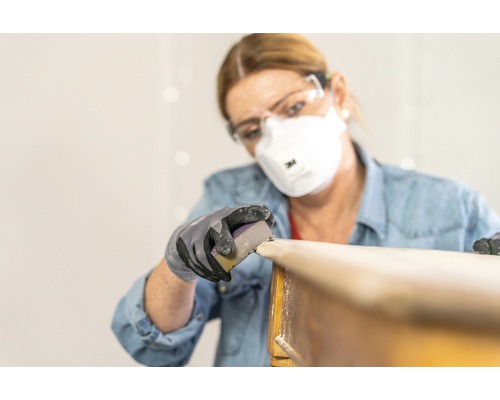 Vrouw schuurt hout met schuurpapier, veiligheidsbril en ademhalingsmasker