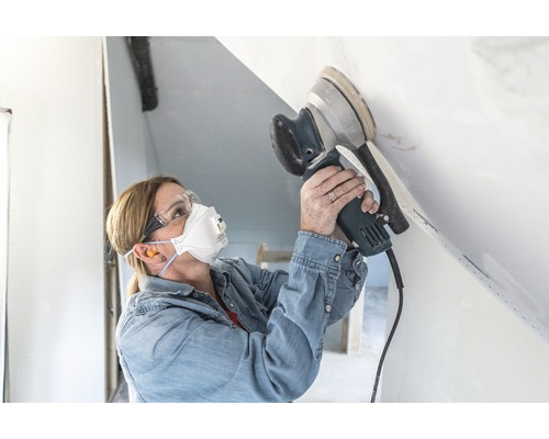 Een vrouw schuurt een plafond met een excentrische schuurmachine en draagt een veiligheidsmasker en een veiligheidsbril.