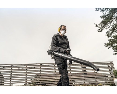 Een persoon bedient een ruggedragen bladblazer in de tuin, beschermd met gehoorbescherming en een stofmasker.