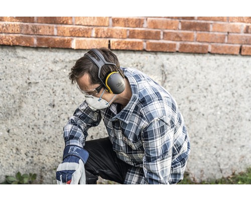 Een man knielt en draagt persoonlijke beschermingsmiddelen, waaronder gehoorbescherming, een veiligheidsbril, een stofmasker en werkhandschoenen.