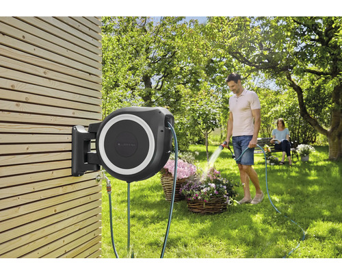Tuinscène met aan een houten wand gemonteerde slangbox en man die bloemen water geeft
