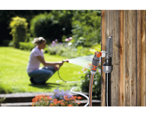 Tuinscène met slangconnector en kraanaansluiting op een houten wand, een vrouw besproeit de tuin