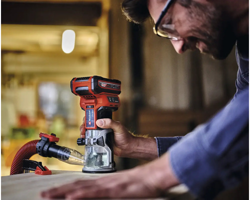 Een man werkt met een Einhell accu bovenfrees op een stuk hout. Er is een afzuiginstallatie aangebracht.