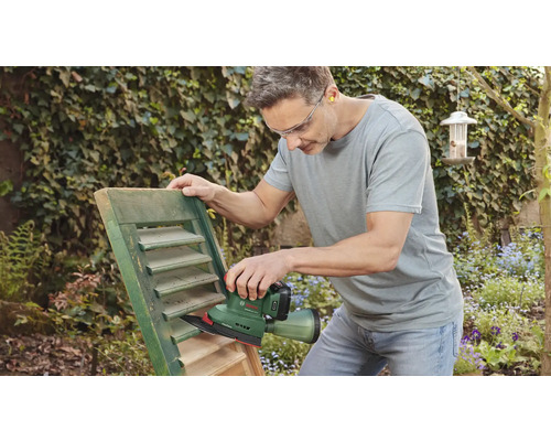 Een man schuurt een luik met een schuurmachine in de tuin.