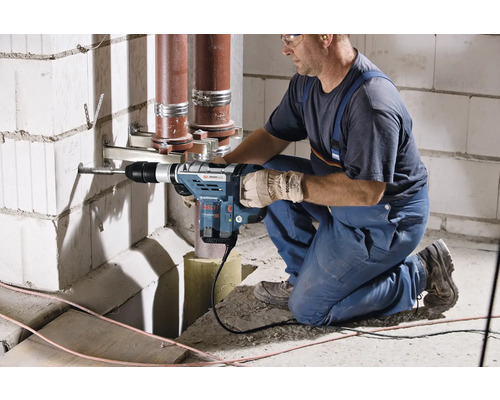 Een vakman boort met een boorhamer een gat in een muur, leidingen lopen op de achtergrond.