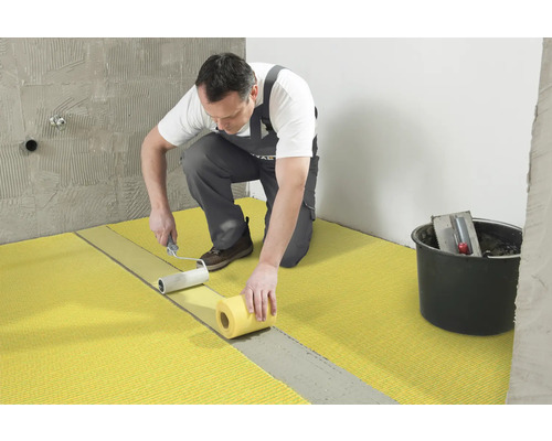 Een vakman brengt met een verfroller een afdichtingsmembraan op de vloer aan. Op de achtergrond staat een emmer met gereedschap.