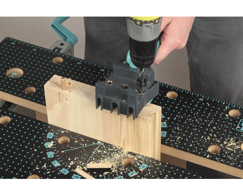 Iemand boort met een boormachine een gat in een stuk hout met een deuvelhulpstuk op een werkbank.