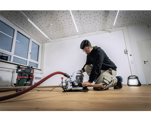 Een man werkt met een Metabo verstekzaag in een lichte ruimte. Op de achtergrond staan een stofzuiger en een bouwlamp.