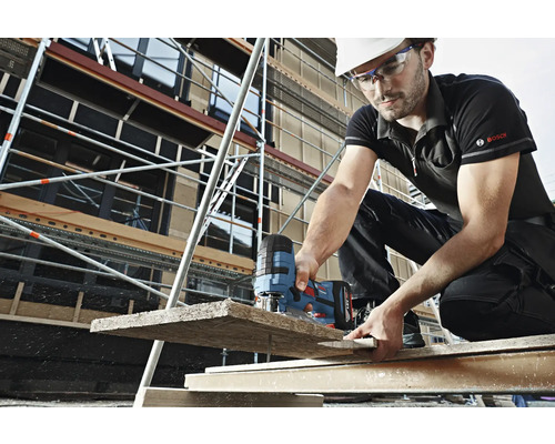 Een bouwvakker met helm en veiligheidsbril zaagt met een decoupeerzaag een houten plank op een bouwplaats. Op de achtergrond is een steiger te zien. Bosch Logo.