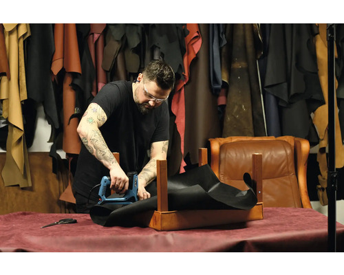 Een vakman bevestigt met een elektrische tacker een zwart stuk stof aan een houten frame in een werkplaats.