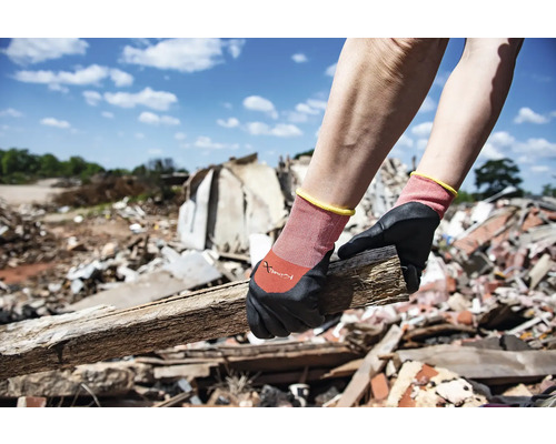Een persoon draagt werkhandschoenen en houdt een stuk hout vast tegen een achtergrond van bouwafval.