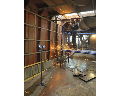 Een arbeider staat op een steiger en snijdt metaal met een snijmachine, waarbij vonken vrijkomen.