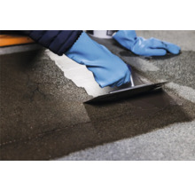 Bitumen daklak wordt aangebracht met een plamuurmes door iemand met handschoenen.