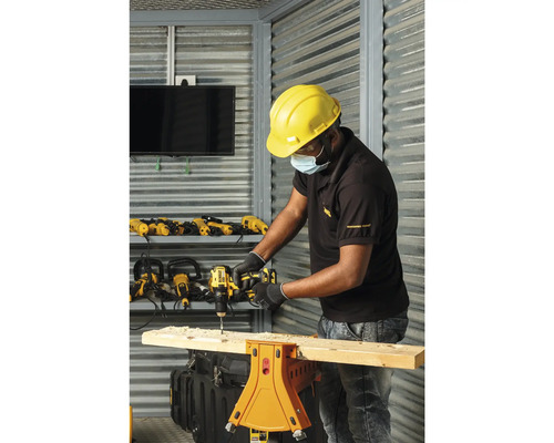 Een man boort met een boormachine een gat in een houten plank die op een werkbank ligt. Op de achtergrond zijn andere boormachines en gereedschappen te zien.