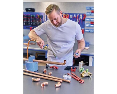 Een man soldeert een koperen buis met een soldeerbrander, terwijl hij soldeertin vasthoudt. Diverse gereedschappen en koperen buisfittingen liggen op tafel.