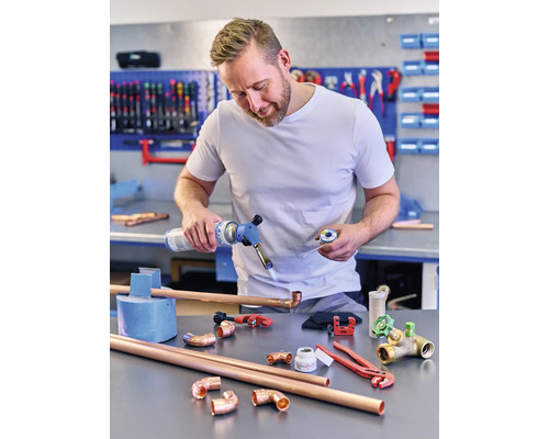 Man soldeert koperen buizen met een gasbrander, omgeven door koperen buisleidingen en gereedschap