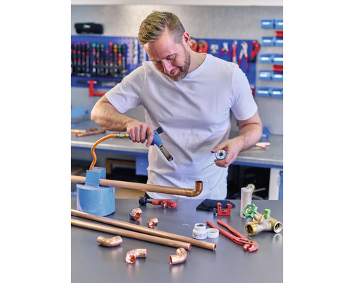 Een man soldeert koperen buizen met een soldeerbout in een werkplaats. Verschillende gereedschappen en buizen liggen op de tafel.