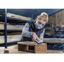 Vrouw schuurt houten kist met schuurblok. Ze draagt een veiligheidsbril en een ademhalingsmasker.