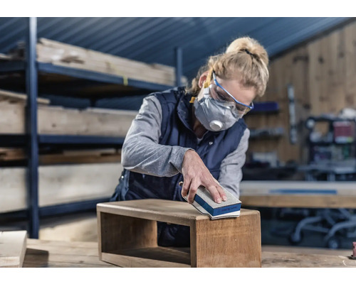 Vrouw schuurt houten kist met schuurblok. Ze draagt een veiligheidsbril en een ademhalingsmasker.