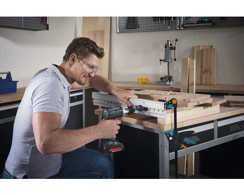 Man boort met een boormachine door een houten plaat met boorsjabloon op een werkbank.