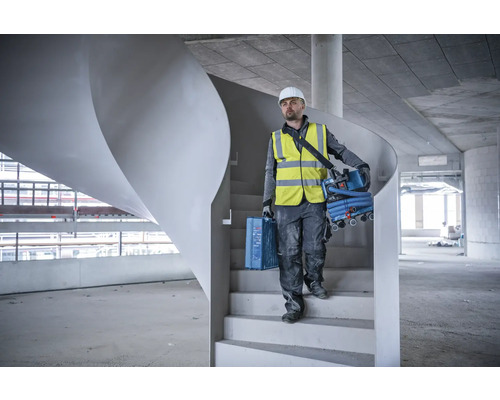 Een bouwvakker met helm en veiligheidsvest draagt een gereedschapskist en een stofzuiger de trap af.