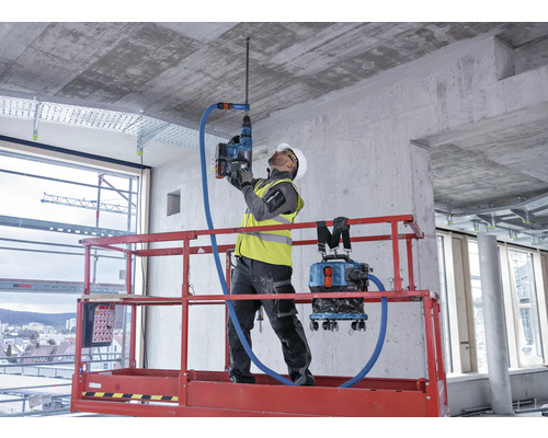 Een bouwvakker boort met een boorhamer een gat in een betonnen plafond. Een stofzuiger is met de boorhamer verbonden om stof te verwijderen.