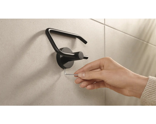 Een hand monteert een zwarte toiletpapierhouder met een inbussleutel aan een wand.