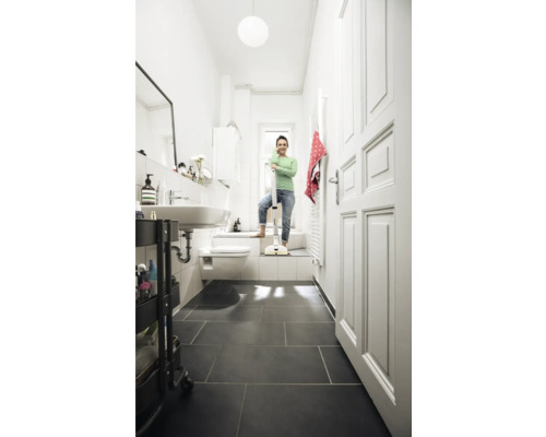 Lichte badkamer met een vrouw die leunt op een draadloze vloerreiniger, wastafel, toilet en donkere tegels.