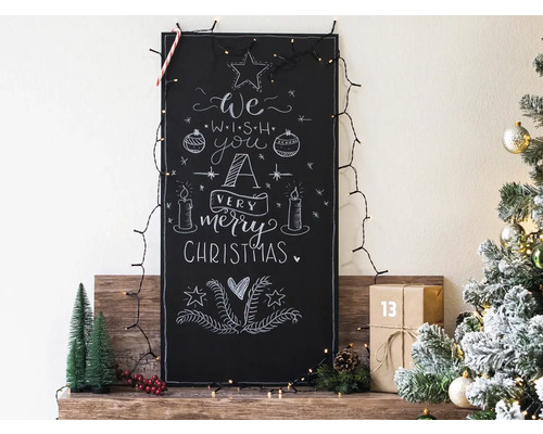 Decoratief bord met krijttekening en kerstverlichting op een houten plank, naast een kerstboom met versiering.