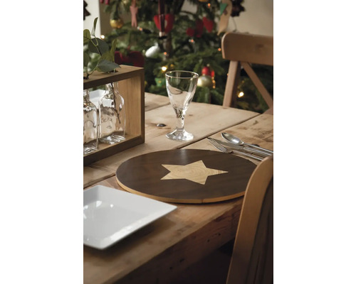 Feestelijk gedekte eettafel met placemat met sterpatroon, glas, bestek en kerstboom op de achtergrond.