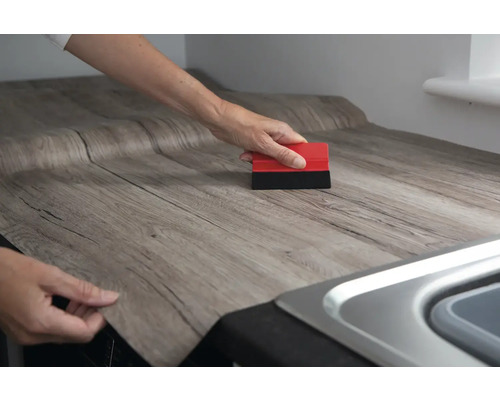 Iemand brengt zelfklevende houtlookfolie aan op een werkblad en gebruikt een rakel.