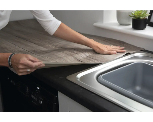 Keukenscène met persoon die zelfklevende decoratiefolie in houtlook op een werkblad aanbrengt.