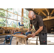 Een man bewerkt een houten werkstuk met een multifunctioneel gereedschap en stofzuiger in een werkplaats.