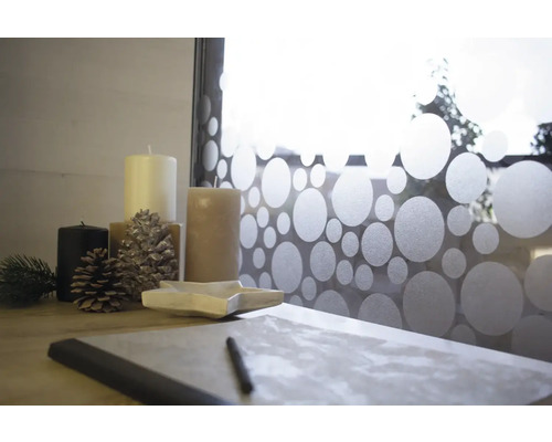 Decoratieve opstelling met kaarsen, dennenappels, stervormige schaal en notitieboek op een tafel voor een raam met stippenpatroonfolie.