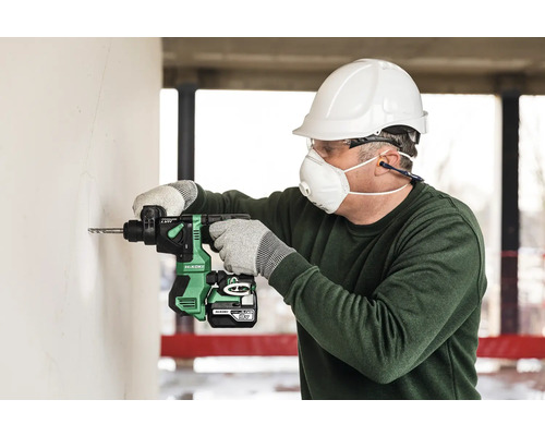 Een man boort met een accuboormachine een gat in een muur, terwijl hij beschermende kleding draagt zoals een helm, bril en masker.