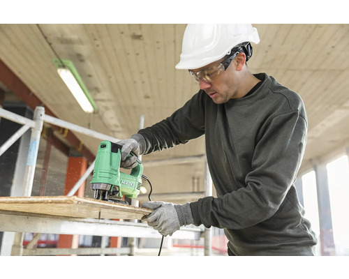 Een man zaagt een houten plank met een decoupeerzaag van het merk Hikoki, terwijl hij beschermende kleding draagt.