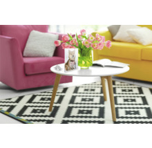 Woonkamer met een witte tafel, bloemen, een foto met een hond, een roze fauteuil, een gele bank en een tapijt met geometrisch patroon.