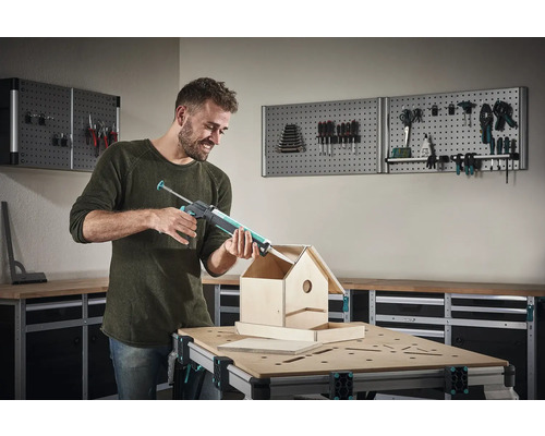 Man gebruikt een kitpistool om een vogelhuisje te bouwen op een werkbank voor een gereedschapswand.