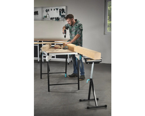 Man boort met een accuboormachine in een stuk hout op een werktafel, op de achtergrond gereedschap aan een gereedschapswand