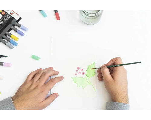 Persoon schildert hulstbladeren met aquarelverf op papier. Aquarelpotloden liggen ernaast.