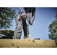 Een persoon boort met een accuboormachine een gat in een houten plank, houtsplinters vliegen.