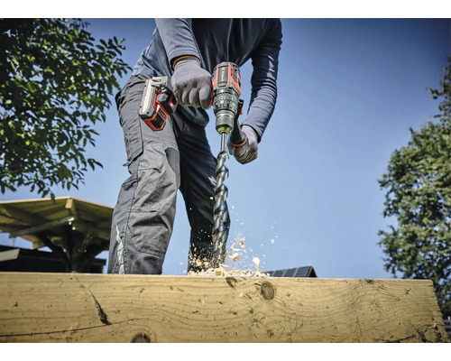 Een persoon boort met een accuboormachine een gat in een houten plank, houtsplinters vliegen.