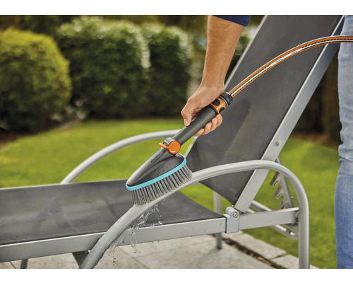 Een persoon reinigt een tuinligstoel met een borstel en een waterslang.
