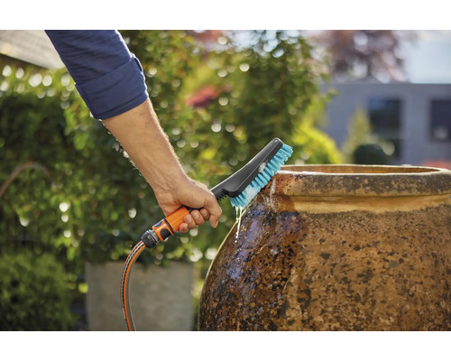 Een persoon reinigt een pot met een borstel die is aangesloten op een waterslang in de tuin.